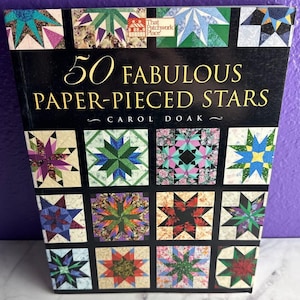 May include: A softcover book titled 50 Fabulous Paper-Pieced Stars by Carol Doak. The black cover features a grid of various colorful star-shaped quilt block patterns in shades of green, blue, pink, red, and purple, with a small logo at the top.