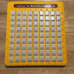 May include: A yellow plastic multiplication learning board titled MagicMath with a grid of ninety-nine individual white square buttons. Each button is printed with a unique multiplication equation ranging from 1x1 to 9x9 in black text.