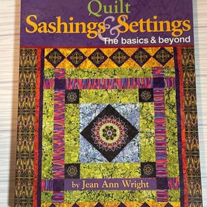 May include: A softcover instructional book titled Quilt Sashings & Settings: The basics & beyond by Jean Ann Wright. The cover features a colorful quilt design with a central mandala pattern, lime green, purple, and orange geometric borders.