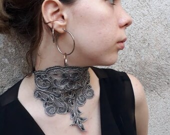 May include: A person wearing a black choker necklace with a delicate lace design. The choker is made of black lace and has a floral pattern. The person is wearing a black top and has their hair pulled back.