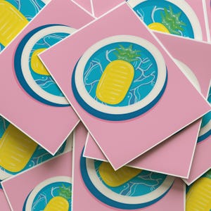 以下が含まれることがあります： ピンクのカードの山で、各カードには青と白の円が描かれています。円の中には、緑の葉が付いた黄色のパイナップルフロートがあります。