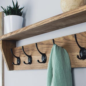 May include: Wooden coat rack with six black metal hooks, mounted on a wall. The rack has a natural wood finish and a shelf above. A green towel hangs from one of the hooks. A plant in a white pot sits on the shelf.