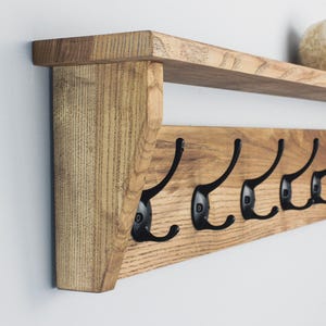 May include: Wooden wall-mounted shelf with seven black metal coat hooks. The shelf is made of light-colored wood and has a simple, rustic design. A decorative item sits on the shelf. The shelf is mounted on a white wall.