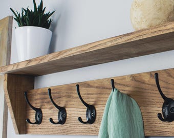May include: A wooden wall-mounted shelf with a lower row of six black metal hooks. A light green towel hangs from one hook. The shelf above holds a white potted plant, a beige sphere, and a textured vase.