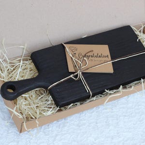 Puede incluir: Una tabla de cortar de madera teñida oscura con un asa, presentada en una caja de cartón con paja decorativa. Una pequeña tarjeta atada con hilo dice "Felicidades con amor para ti."