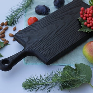 Op de afbeelding: Een zwart houten snijplank met een handvat, omringd door vers fruit en noten. De plank heeft een getextureerd oppervlak en een gat aan het einde van het handvat. Peren, pruimen, amandelen en rode bessen zijn rond de plank gerangschikt.