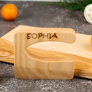 Könnte beinhalten: Ein helles Holzschneidebrett mit einem markanten Griff, auf dem der Name "SOPHIA" eingebrannt ist. Im Hintergrund sind zwei Orangen und ein dekorativer Ball zu sehen.