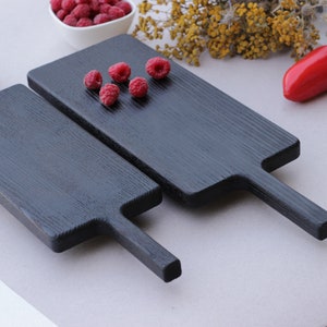 Op de afbeelding: Twee zwarte houten serveerplanken met handvatten worden op een licht oppervlak getoond. Verse frambozen zijn op een plank geplaatst. Rode paprika's en een kom met frambozen staan ook in de afbeelding. De planken hebben een rechthoekige vorm.