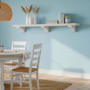 May include: A white wooden wall-mounted shelf with three decorative corbel supports. The shelf holds three white vases with dried wheat stalks, a small stack of books, and a small succulent plant in a blue and white striped ceramic pot.