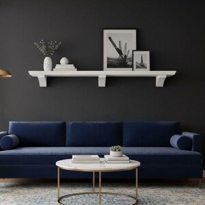 May include: A contemporary living room featuring a navy blue sofa, a white armchair, and a round coffee table with a gold base. A patterned rug and a gold floor lamp enhance the decor. The walls are dark grey with a white shelf and framed art.