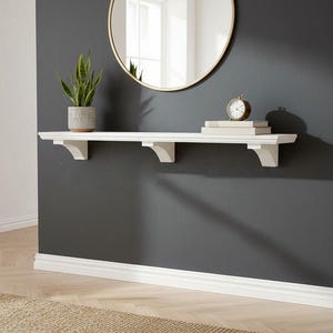 May include: A long white wall-mounted shelf featuring three decorative corbel brackets. The shelf is positioned horizontally and holds a potted snake plant, a small analogue clock, and a stack of books, providing a functional storage surface.