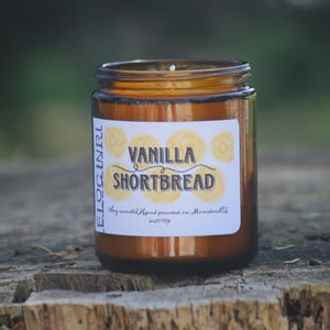 May include: A brown glass jar candle with a white label that reads "Vanilla Shortbread" in a bold font. The label also features yellow circular designs. The candle is set on a wooden surface.