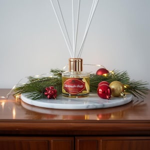 May include: A clear glass reed diffuser bottle with a gold-toned cap and white reeds. The label features red and gold accents with the text "Christmas in a Bottle". The bottle is surrounded by pine branches, red and gold ornaments, and string lights.
