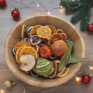 May include: A large, round, light-coloured wooden bowl filled with a variety of dried fruit slices, including orange, lime, and apple, along with a small orange pumpkin-shaped gourd and green leaves, creating a festive, natural holiday display.