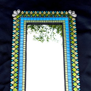 May include: A rectangular mirror with a mosaic frame. The frame is made of small, square tiles in shades of blue, green, and gold. The tiles are arranged in a geometric pattern. The mirror is surrounded by a decorative border of gold leaf and green glass.