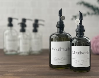 May include: Two amber glass bottles with black pump tops and white labels that read "Mouthwash Signature Collection" with the size in ounces. The bottles are on a wooden surface.