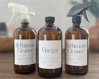 May include: Three amber glass spray bottles with white labels. The labels have black text that reads "All Purpose Cleaner", "Vinegar", and "Bathroom Cleaner". Each bottle has a black spray nozzle and a white label that says "Signature Collection" and "16 ounces".