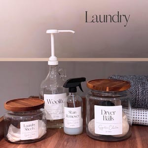 May include: A collection of laundry essentials, including glass jars with wooden lids, a spray bottle, and a dispenser bottle. The jars are labeled "Laundry Sheets" and "Dryer Balls." The spray bottle is labeled "Stain Remover."