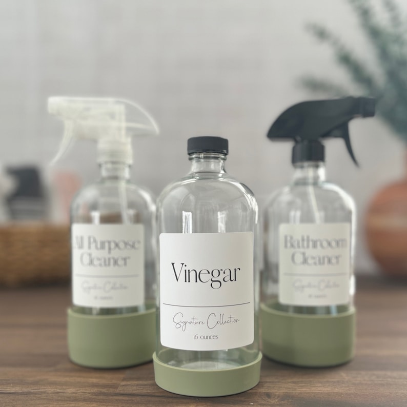Op de afbeelding: Drie transparante glazen spuitflessen met zwarte en witte etiketten. De flessen zijn gelabeld met "All Purpose Cleaner", "Vinegar Signature Collection 16 ounces" en "Bathroom Cleaner". De flessen staan op een houten oppervlak.