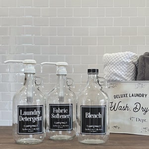 May include: Three glass bottles with black labels and white lettering. The bottles are labeled "Laundry Detergent", "Fabric Softener", and "Bleach". The bottles are sitting on a counter with a white tile backsplash. A white metal sign with black lettering reads "DELUXE LAUNDRY Wash. Dry 47&deg; Dept."