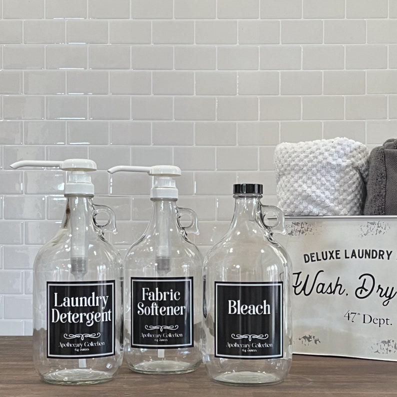 May include: Three glass bottles with black labels and white lettering. The bottles are labeled "Laundry Detergent", "Fabric Softener", and "Bleach". The bottles are sitting on a counter with a white tile backsplash. A white metal sign with black lettering reads "DELUXE LAUNDRY Wash. Dry 47&deg; Dept."