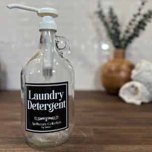 May include: A clear glass bottle with a white pump dispenser and a black label that reads "Laundry Detergent" with the text "Apothecary Collection 64 ounces" below.