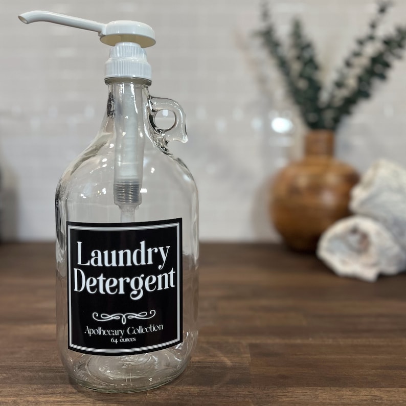 May include: A clear glass bottle with a white pump dispenser and a black label that reads "Laundry Detergent" with the text "Apothecary Collection 64 ounces" below.