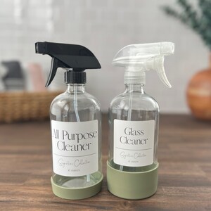 Op de afbeelding: Twee transparante glazen spuitflessen met zwarte en transparante spuitkoppen. De flessen hebben groene siliconen bases en etiketten met de tekst "All Purpose Cleaner" en "Glass Cleaner".