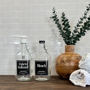 May include: Two glass bottles with black labels and white text. The bottle on the left says "Fabric Softener" and the bottle on the right says "Bleach". Both bottles have a black and white label with the text "Apothecary Collection".