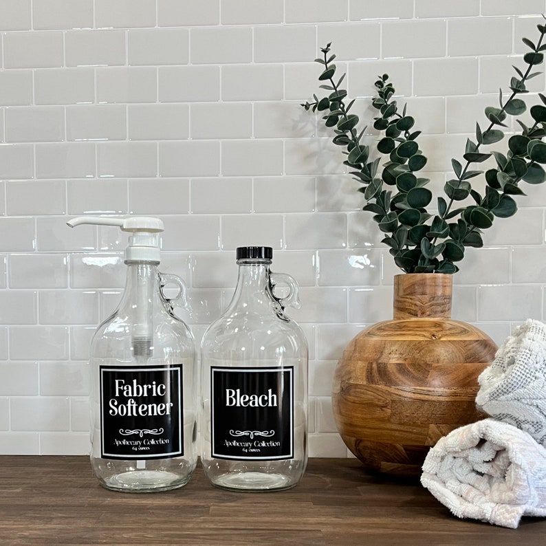 May include: Two glass bottles with black labels and white text. The bottle on the left says "Fabric Softener" and the bottle on the right says "Bleach". Both bottles have a black and white label with the text "Apothecary Collection".