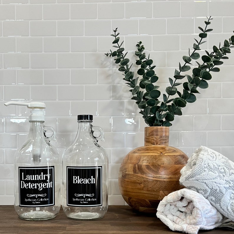 May include: Two glass bottles with black labels and white text. The labels read "Laundry Detergent" and "Bleach". The bottles are on a wooden countertop with a wooden vase with green leaves and white towels.