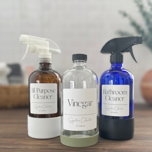 Op de afbeelding: Drie glazen spuitflessen met etiketten. De flessen zijn gelabeld met "All Purpose Cleaner", "Vinegar" en "Bathroom Cleaner". Elke fles heeft een zwarte spuitmond en een wit etiket met zwarte tekst. De flessen staan op een houten oppervlak.