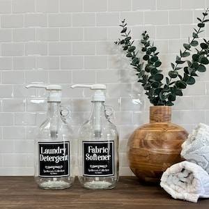 May include: Two clear glass bottles with white pump tops and black labels. The labels read "Laundry Detergent" and "Fabric Softener". The bottles are sitting on a wooden surface in front of a white tiled wall.