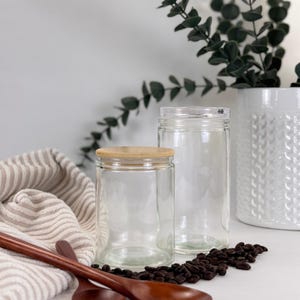 May include: Two clear glass jars, one with a wooden lid, sit on a white surface. Coffee beans are scattered around the jars. A striped cloth and wooden spoons are in the foreground. A white vase with greenery is in the background.
