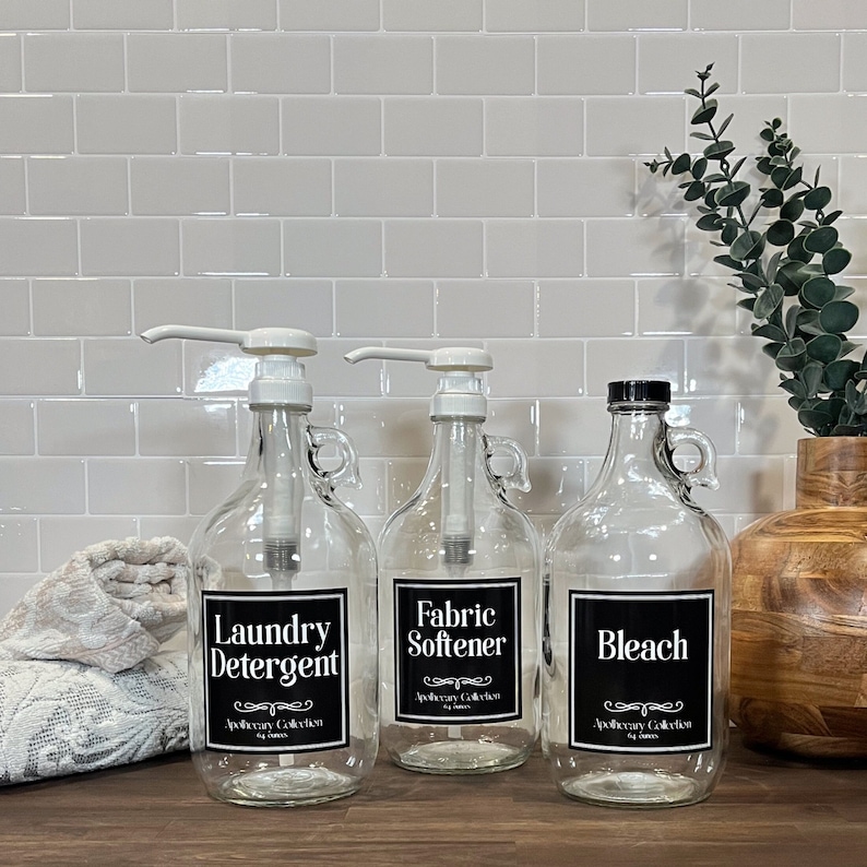 May include: Three glass bottles with pump tops and labels. The bottles are labeled "Laundry Detergent", "Fabric Softener", and "Bleach". The bottles are on a wooden surface with a white tiled background.