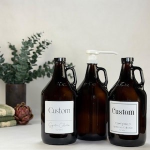 May include: Three amber glass jugs with handles and black screw-top lids. Two jugs feature white rectangular labels with black text reading Custom, Signature Collection, and Apothecary Collection. One jug has a white plastic pump dispenser.