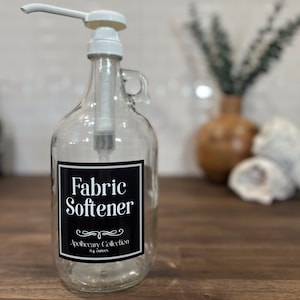 May include: A clear glass bottle with a white pump dispenser and a black label that reads "Fabric Softener" with the text "Apothecary Collection 64 ounces" below.