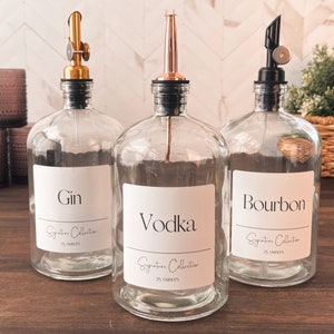May include: Three clear glass bottles with black, gold, and copper spouts. Each bottle has a white label with black text. The labels read "Gin", "Vodka", and "Bourbon". Each bottle also has a label that reads "Signature Collection 25 ounces".