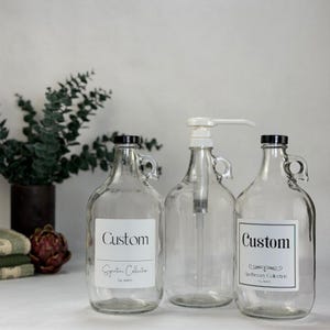 May include: Three clear glass jugs with handles, each featuring a black cap. Two jugs have white labels with the text Custom and Signature Collection or Apothecary Collection. The center jug includes a white pump dispenser for liquid contents.