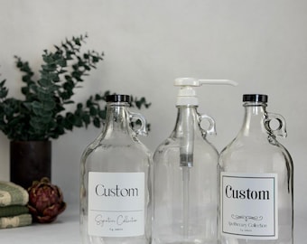 May include: Three clear glass jugs with handles, each featuring a black cap. Two jugs have white labels with the text Custom and Signature Collection or Apothecary Collection. The center jug includes a white pump dispenser for liquid contents.