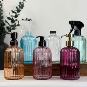 May include: A selection of decorative glass bottles in shades of amber, pink, clear, and teal. Some bottles feature pump dispensers, while others have spray nozzles. The bottles are displayed on a wooden surface.