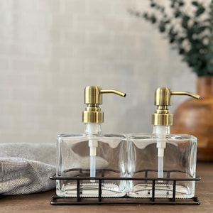 May include: Two clear glass soap dispensers with gold pumps, sitting in a black metal wire rack. The dispensers are square shaped and have a modern design.