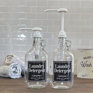 May include: Two clear glass bottles with black labels that say "Laundry Detergent" and "Apothecary Collection" on a white tiled countertop. The bottles have white pump tops and are filled with a clear liquid.