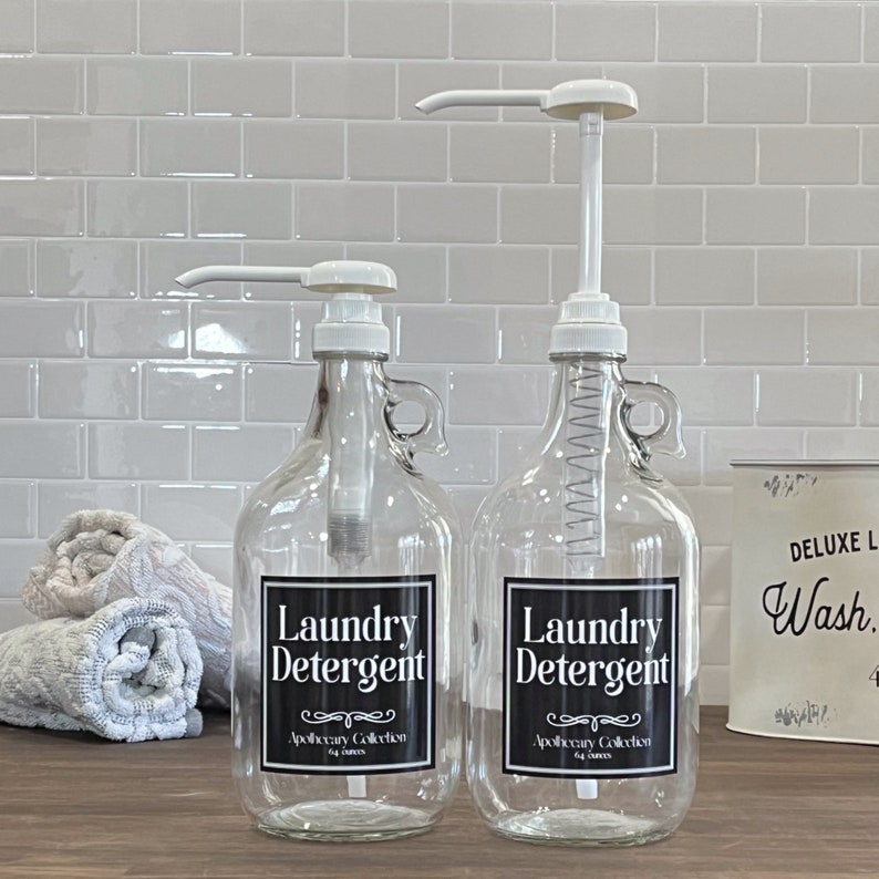 May include: Two clear glass bottles with black labels that say "Laundry Detergent" and "Apothecary Collection" on a white tiled countertop. The bottles have white pump tops and are filled with a clear liquid.