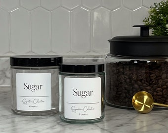 May include: Two glass jars with black lids and white labels that say "Sugar Signature Collection" with the size in ounces. The larger jar is 16 ounces and the smaller jar is 8 ounces. The jars are sitting on a white marble countertop.