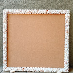 May include: A corkboard with a white seashell frame. The frame is made of small, white seashells that are glued together to form a border around the corkboard.