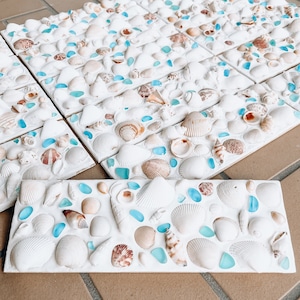 May include: A close-up of a white mosaic tile with seashells and blue glass pieces. The tile is made of a smooth, white material and features a variety of seashells in different sizes and shapes. The blue glass pieces are scattered throughout the tile, adding a touch of color and sparkle.
