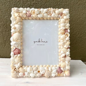 May include: A rectangular photo frame decorated with various seashells in shades of white, cream, and pink. The frame has the text "park lane PHOTO FRAME (8x10)" in the lower right corner.