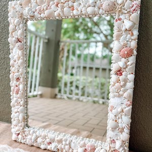 May include: A white mirror with a decorative frame made of seashells. The frame is made of a variety of seashells, including white, pink, and brown shells. The mirror is reflecting a blurry image of a patio.
