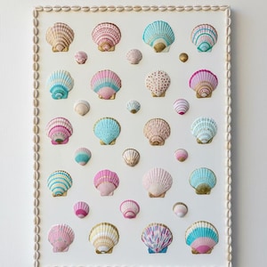 May include: A framed artwork featuring a collection of painted seashells in various sizes and colors, including pink, blue, and gold. The shells are arranged on a white background and framed with small seashells.